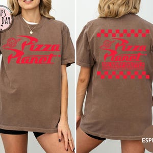 May include: A brown t-shirt with the Pizza Planet logo in red on the front and back. The front logo includes a pizza slice graphic, and the back has a red and white checkered pattern. The shirt is a casual, unisex style, in a brown colour.