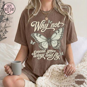 May include: A brown short-sleeve t-shirt featuring a large graphic of a butterfly in the center. The text "Why not spread your wings and fly" is printed in a cream-colored script font above and below the butterfly. The shirt has a crew neckline.