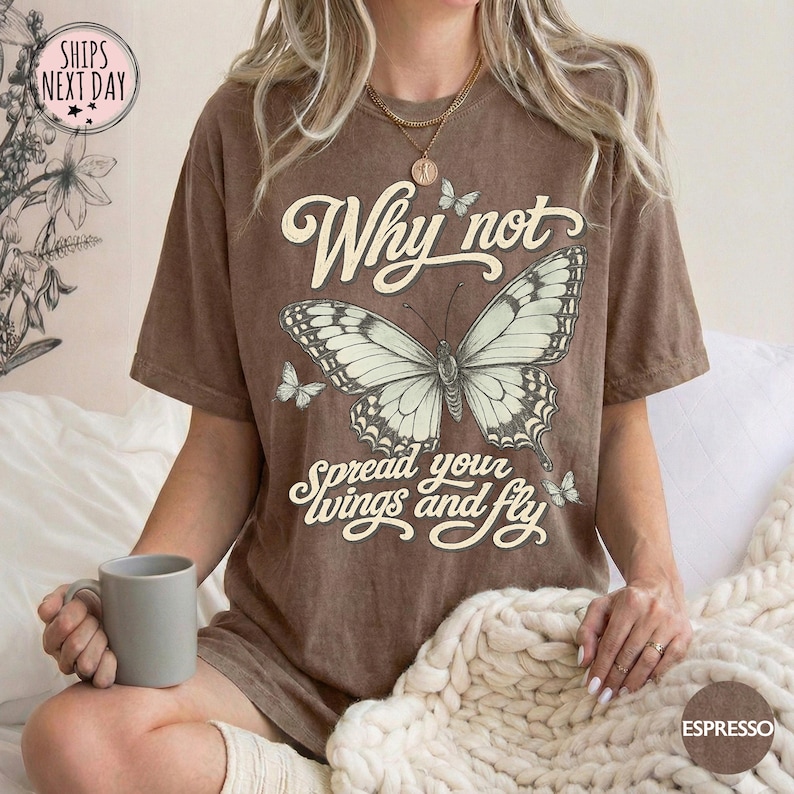 May include: A brown short-sleeve t-shirt featuring a large graphic of a butterfly in the center. The text "Why not spread your wings and fly" is printed in a cream-colored script font above and below the butterfly. The shirt has a crew neckline.