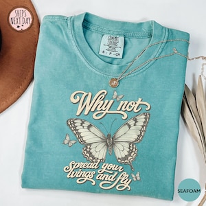 May include: A seafoam green short-sleeve t-shirt featuring a large butterfly graphic in the center. The text reads Why not spread your wings and fly in a cursive font. The shirt has a crew neckline and a Comfort Colors brand label at the neck.