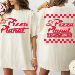 May include: Ivory t-shirt featuring the red "Pizza Planet" logo on the front and back. The back design includes "DELIVERY SHUTTLE" and "SERVING YOUR LOCAL STAR CLUSTER" text, with a red and white checkered pattern.