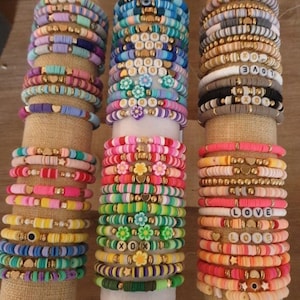May include: A display of colorful beaded bracelets. The bracelets feature various colors, including blue, pink, yellow, and green, with gold accents and letter beads spelling out words like "LOVE" and "MOM". Some bracelets have flower and heart charms.