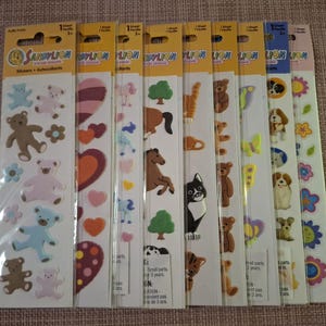 Puede incluir: Una colección de hojas de pegatinas Sandy Lion con varios diseños. Las pegatinas incluyen osos de peluche, corazones, flores, caballos, gatos, perros y mariposas. Cada hoja está empaquetada en una funda de plástico transparente con un encabezado amarillo.
