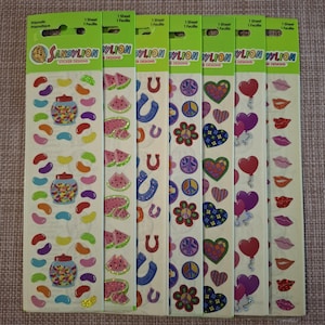 Puede incluir: Una colección de hojas de pegatinas Sandylion con varios diseños. Las hojas muestran imágenes coloridas de gominolas, sandías, herraduras, símbolos de la paz, corazones, globos y labios. Cada hoja está etiquetada con el texto "1 Sheet" y "Prismatique".