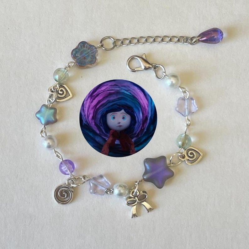 May include: A silver-toned charm bracelet featuring a round image of a doll with a purple and blue background. The bracelet includes star, heart, and bow charms, along with iridescent and purple beads. The bracelet has a lobster clasp closure.
