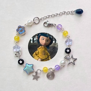 May include: A silver-toned bracelet with a variety of beads and charms. The bracelet features yellow, purple, and clear beads, along with star and flower charms. A small image of a character in a yellow coat is in the center.