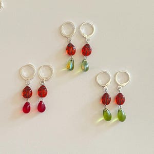 May include: Three pairs of silver hoop earrings, each featuring a red ladybug charm with black spots. One pair has red teardrop beads, while the other two pairs feature translucent green teardrop beads hanging below the ladybug charms.