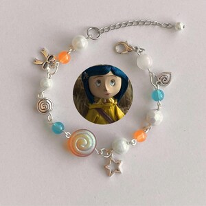May include: A silver-toned bracelet featuring a Coraline doll charm, adorned with white, orange, and blue beads. The bracelet includes a star, bow, and swirl charms. The adjustable chain measures approximately 7 inches.