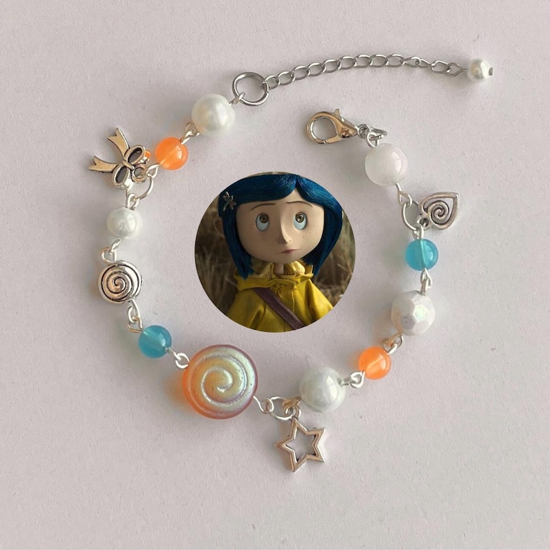 May include: A silver-toned bracelet featuring a Coraline doll charm, adorned with white, orange, and blue beads. The bracelet includes a star, bow, and swirl charms. The adjustable chain measures approximately 7 inches.