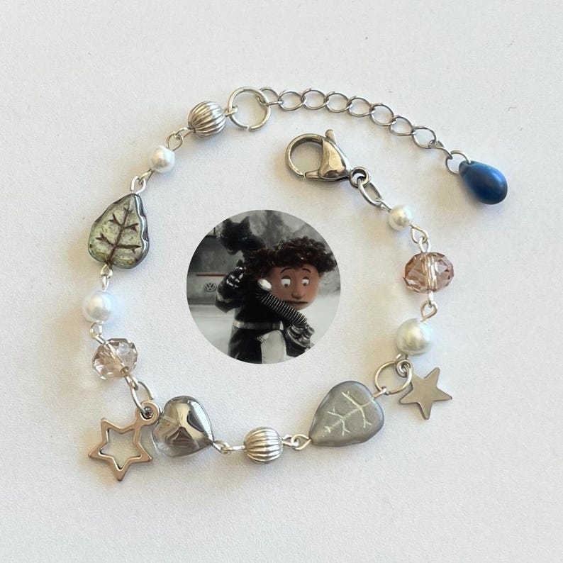 May include: Silver-toned bracelet featuring a variety of beads, including pearls, clear and brown faceted beads, and leaf-shaped and heart-shaped charms. The bracelet also includes a star charm and a blue teardrop bead. A small circular image of a cartoon character is in the center.