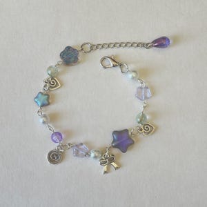 May include: A delicate bracelet featuring a mix of iridescent beads in shades of purple, blue, and green. The bracelet includes star, flower, and heart-shaped charms, along with a silver-toned bow charm. The adjustable chain ends with a teardrop-shaped bead.