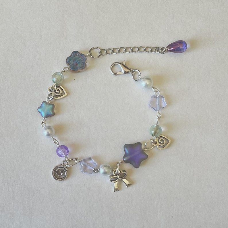May include: A delicate bracelet featuring a mix of iridescent beads in shades of purple, blue, and green. The bracelet includes star, flower, and heart-shaped charms, along with a silver-toned bow charm. The adjustable chain ends with a teardrop-shaped bead.