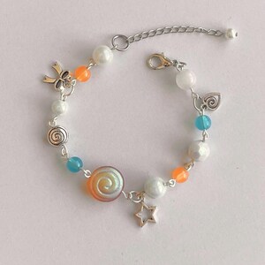 May include: A beaded bracelet with a silver chain and clasp. The bracelet features white, orange, and blue beads, along with silver charms including a bow, a star, and a spiral design. The bracelet is approximately 7 inches long.