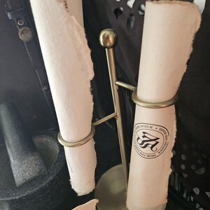 May include: A brass scroll holder with two rolled parchment scrolls. One scroll has a circular emblem with the words "Witch" and "Written From The Light". The parchment has a rough, torn edge.