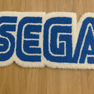 Może przedstawiać: Niebiesko-biały dywan z napisem "SEGA" w odważnej, stylizowanej czcionce. Litery są niebieskie z białym obramowaniem, na białym tle. Dywan ma miękki, teksturowany wygląd i jest umieszczony na drewnianej powierzchni.