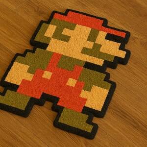 Puede incluir: Una alfombra pixelada con el personaje de videojuego Mario en una pose de carrera. La alfombra es principalmente roja, naranja, amarilla, verde y negra, con un borde negro. La alfombra está colocada sobre un suelo de madera.