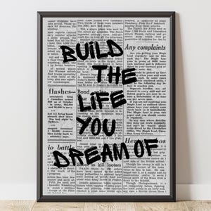 Könnte beinhalten: Schwarz gerahmter Druck mit einem Vintage-Zeitungs-Hintergrund. Der Text-Overlay lautet "BUILD THE LIFE YOU DREAM OF". Der Druck hat ein motivierendes Thema mit Retro-Ästhetik.