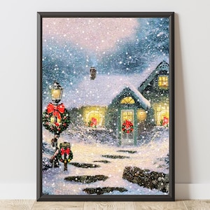 Könnte beinhalten: Gerahmter Druck einer verschneiten Winterszene. Ein reizendes Häuschen mit beleuchteten Fenstern und einem Kranz an der Tür ist von fallendem Schnee umgeben. Eine Straßenlaterne mit roter Schleife und Kranz steht im Vordergrund.