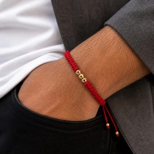 May include: A red woven cord bracelet featuring three small, polished gold-tone spherical beads centered on the band. The bracelet includes adjustable red cord ends, each finished with a single small gold-tone bead at the tip for a secure fit.