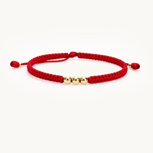 May include: Red macrame bracelet with three gold-colored beads. The adjustable bracelet features a sliding knot closure and small gold-colored accents at the ends. A simple, elegant piece of jewelry.