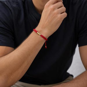 May include: Red braided bracelet with three gold beads. The bracelet is tied with a sliding knot for adjustable sizing. The bracelet is worn on a wrist.