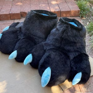 May include: A pair of large, black furry paw-shaped slippers with light blue claws. The slippers have a plush, textured appearance and are designed to resemble animal paws. The interior of the slippers is a light blue color.