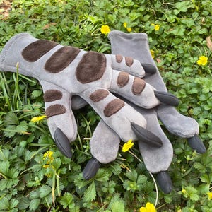 May include: A pair of grey gloves with brown spotted accents and black claw-like tips. The gloves are designed to resemble a creature's paws and are lying on a bed of green grass with yellow flowers.