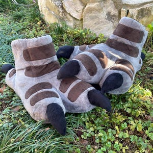 Puede incluir: Un par de zapatillas de felpa con forma de pies de monstruo. Las zapatillas son grises con detalles marrones y grandes garras negras. Están diseñadas para parecerse a los pies de un monstruo y están sobre hierba verde.