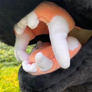 May include: Close-up of a plush toy's open mouth, revealing soft, white teeth and a peach-colored interior. The outer part of the toy is black and furry, with a focus on the detailed stitching and texture.