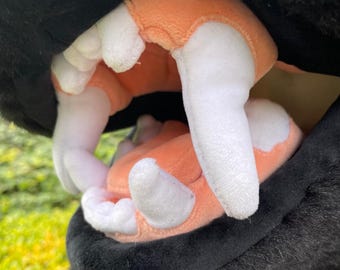May include: Close-up of a plush toy's open mouth, revealing soft, white teeth and a peach-colored interior. The outer part of the toy is black and furry, with a focus on the detailed stitching and texture.