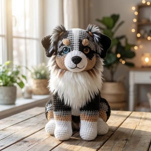 Puede incluir: Un juguete de perro tejido a ganchillo con un patrón negro, blanco y marrón. El perro tiene un ojo azul y un ojo marrón, con un cuello blanco esponjoso. Está sentado sobre una superficie de madera.