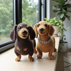 Puede incluir: Dos figuras de perro salchicha de ganchillo. Una es marrón y canela, la otra es canela con un collar de cadena plateada. Están de pie sobre una superficie de madera clara frente a una ventana con vegetación visible afuera.