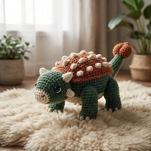 Puede incluir: Un juguete de dinosaurio tejido a mano en tonos verdes, naranjas y crema. El dinosaurio tiene una espalda texturizada, pequeños cuernos y una cola redondeada. Está sobre una alfombra esponjosa de color crema.