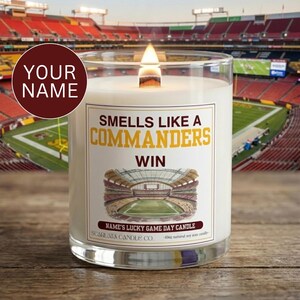 May include: A clear glass candle with a white wax fill and a lit wooden wick. The label features the text "SMELLS LIKE A COMMANDERS WIN" in yellow and brown, an illustration of a stadium, and the text "NAME'S LUCKY GAME DAY CANDLE" and "10oz".