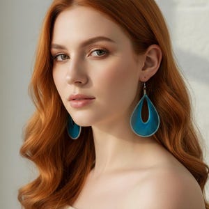 May include: A woman with long, wavy auburn hair wearing teal teardrop earrings. The earrings have a woven texture and silver-coloured trim. She is wearing an off-the-shoulder cream-coloured top.