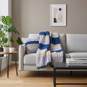 May include: A gray sofa with a blue, white, and purple striped throw blanket and matching pillow. A black side table holds books. A framed abstract art piece hangs on the wall. A potted plant and a coffee cup sit on a small wooden table.