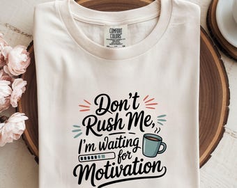 May include: Cream-colored t-shirt with the text "Don't Rush Me, I'm waiting for Motivation" in black script. The design includes a coffee cup and a loading bar graphic. The shirt is made of a soft material.