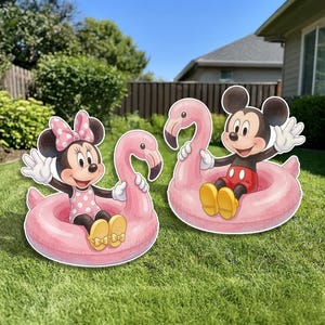 May include: Two decorative yard signs featuring Minnie Mouse and Mickey Mouse, each sitting inside a pink flamingo-shaped pool float. Minnie wears a pink polka-dot dress and bow, while Mickey wears red shorts and yellow shoes, both waving hands.