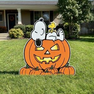May include: A yard sign featuring a white Snoopy character lying on top of a large orange jack-o'-lantern with a glowing yellow face, accompanied by a small yellow Woodstock bird, surrounded by several smaller orange pumpkins at the base.