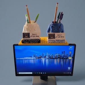 May include: A yellow monitor top shelf holding two 3D-printed pen holders shaped like medical scrubs. The left holder is white with a black nameplate reading Dr. V. Frankenstein Organ Specialist. The right is blue with a nameplate reading Florence Nightingale 1st professional nurse. Both feature Steve's 3D Gift Shop labels.