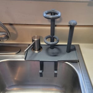 May include: A gray plastic sink organizer with a built-in soap dispenser and holders for a sponge and a brush. The organizer sits on the edge of a stainless steel sink.