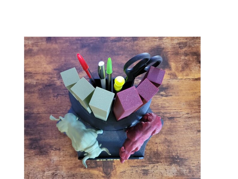K&ouml;nnte beinhalten: Ein schwarzer Schreibtisch-Organizer mit einzigartigem Design, der Stifte, Scheren und farbige Bl&ouml;cke enth&auml;lt. Der Organizer verf&uuml;gt &uuml;ber zwei tierf&ouml;rmige Figuren in Gr&uuml;n und Burgunderrot. Der Schreibtisch-Organizer steht auf einer Holzoberfl&auml;che.