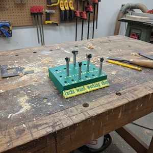 May include: A green wood bit organizer with various drill bits is displayed on a wooden workbench. The organizer has labeled holes for different bit sizes. The text "Jacks wood working shop" is printed on the organizer.