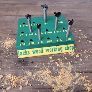 May include: A green rectangular drill bit organizer block with yellow printed fractional inch labels and five metal drill bits inserted. The front yellow base panel features the text Jacks wood working shop in green lettering.