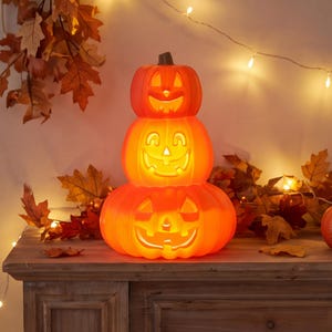 May include: A stack of three orange plastic jack-o'-lanterns with carved faces, illuminated from within to create a warm glow. The pumpkins are arranged vertically, with the smallest on top and the largest on the bottom, featuring cut-out eyes, noses, and wide, toothy grins.