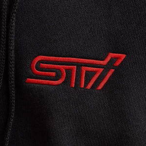 May include: Black hoodie with a red embroidered STI logo on the chest. The logo is a stylized design with the letters "S", "T", and "I" in a bold, modern font. The hoodie has a drawstring and a ribbed hem.