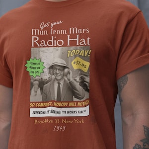 Pode incluir: Camiseta cor ferrugem com um anúncio vintage para um "Radio Hat". O anúncio inclui uma foto em preto e branco de um homem usando o chapéu, com o texto "Ouça música em movimento!" e "Brooklyn 33, Nova York 1949."