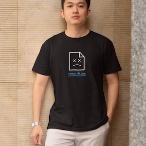 Pode incluir: Camiseta preta com um gráfico branco de um ícone de arquivo com um rosto triste e olhos riscados. O texto "image 01.jpg" está abaixo. A pessoa está vestindo calças cinza claro.
