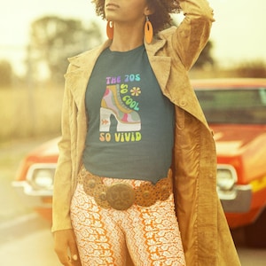 May include: A woman wearing a teal t-shirt with a colorful 70s-themed graphic and the text "THE 70S SO COOL SO VIVID." She is also wearing orange patterned pants, a brown belt, and a tan jacket. The woman is wearing large square sunglasses and orange earrings.