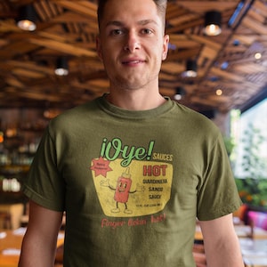 May include: Olive green t-shirt featuring a vintage-style graphic for "¡Oye! Sauces." The design includes a cartoon bottle of hot sauce with outstretched arms, the text "HOT," and "Finger lickin hot!"